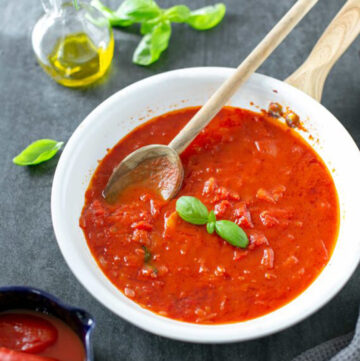 italian tomato sauce in a white pan with a wood spoon, next to a grey napkin on the right side, on the left side a small bowl with canned tomatoes, basil leaves and a small glass bottle of extra-virgin olive oil.
