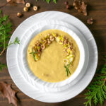 Leek and Potato soup in white bowl over two white plates, spoon on the side over a white napkin. brown and green leaves, and toasted hazelnuts scattered over a wood table.