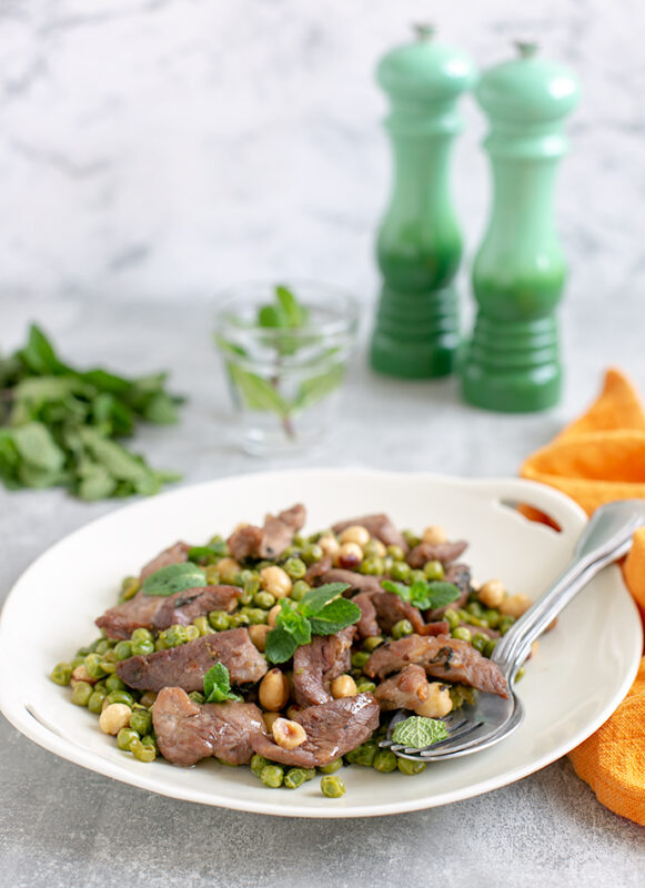 Air Fryer Mint Lamb with Toasted Hazelnut and Peas on a white serving dish, salt and pepper mills in the background together with a glass of water and extra mint leaves.