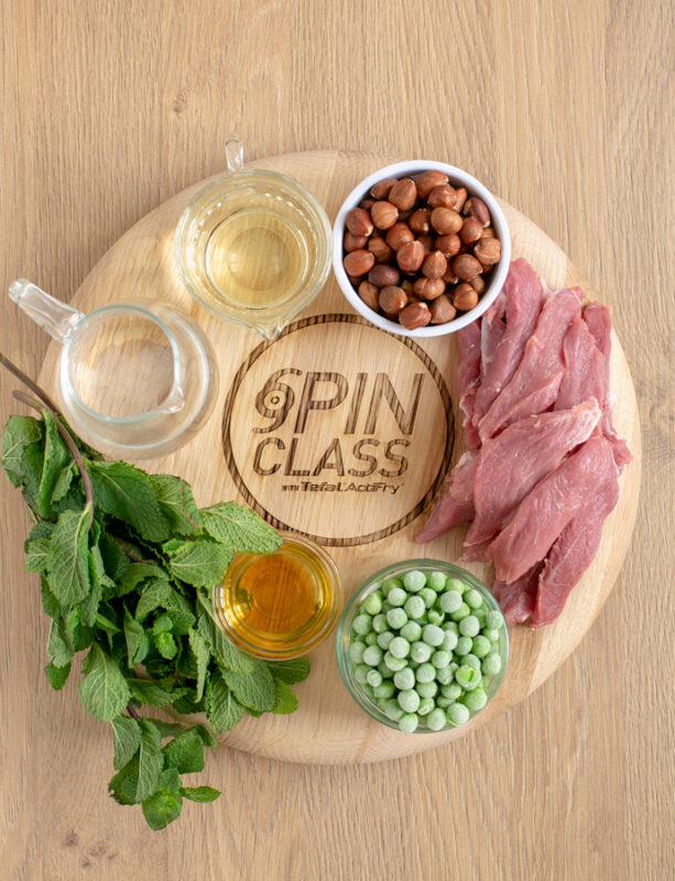 woodboard with fresh mint leaves, hazelnut oil in a small pot, frozen peas in a small pot, strips of lamb meat, hazelnuts in a small pot, white wine in a small pot, and water in a small jug