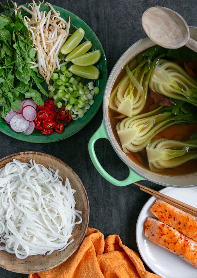 pho broth with shiitake and pak choi in a large pot,  a plate with rice noodles, a plate with grilled salmon, a plate with pho toppings.