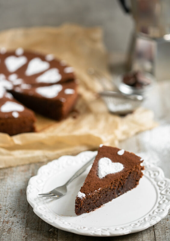 italian caprese cake (gluten-free chocolate cake) slice served on a white plate, whole cake in the background