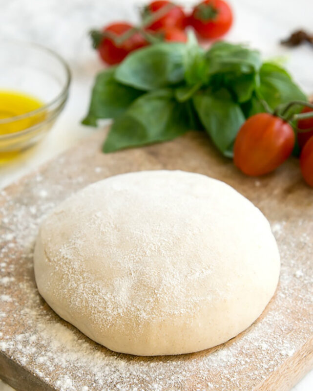 homemade pizza dough on a wood board, tomatoes, basil and olive oil in the background