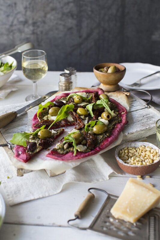 Beet Pizza with Beet Leaf Pesto, topped with capers, olives and sun dried tomatoes for loads of flavour. Shown on a table set for two with wine and salad.