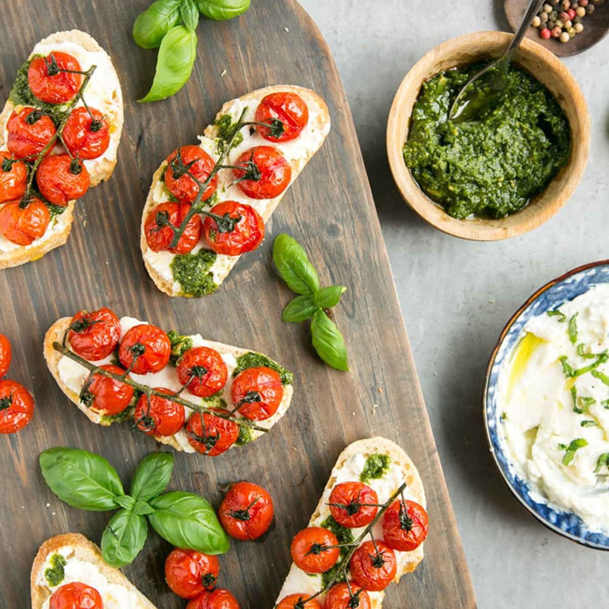 Cherry tomato confit bruschetta with ricotta and basil pesto over wood board, served with ricotta and pesto on the side.