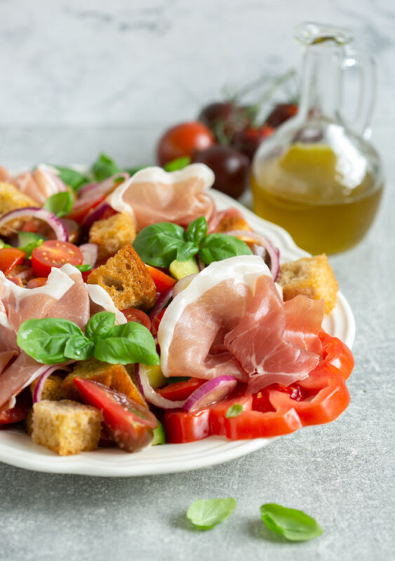 panzanella salad with parma ham on a serving dish, bottle of olive oil and tomatoes in the background
