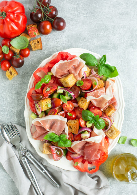 panzanella salad with parma ham on a serving dish, next to mixed tomatoes, bottle of olive oil and napkin with fork and spoon over it