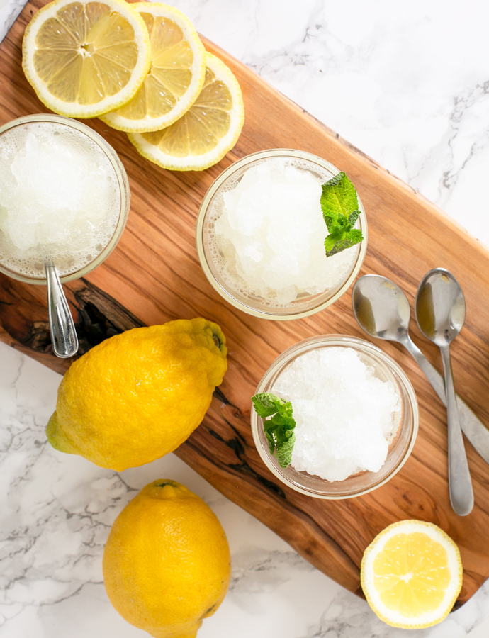 three glasses with lemon granita topped with mint leaves, two lemons, slices of lemon and spoons.