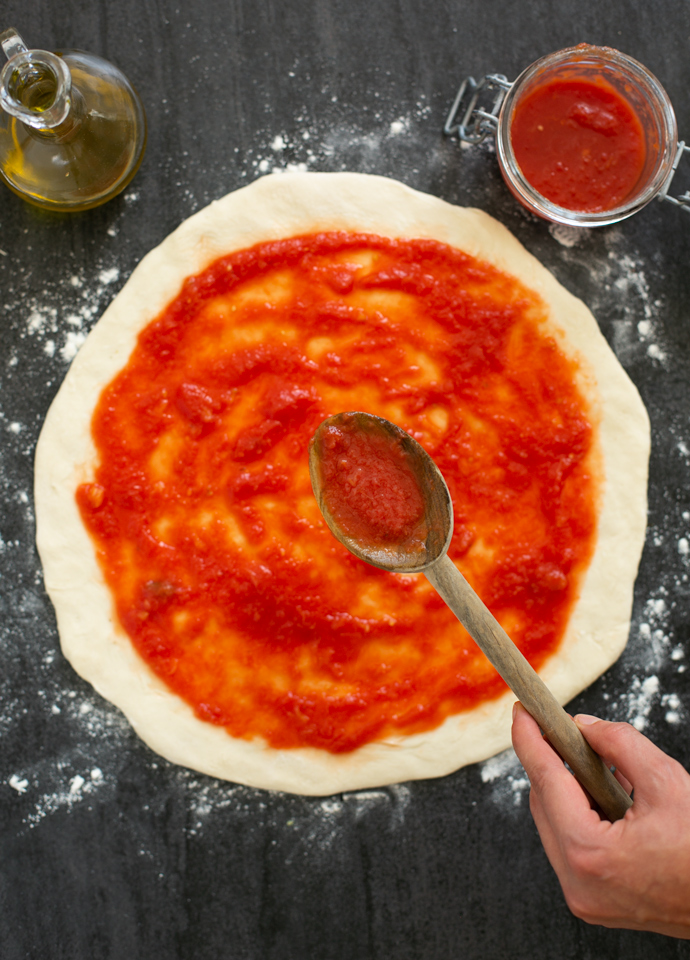 hand spooning pizza sauce on pizza base