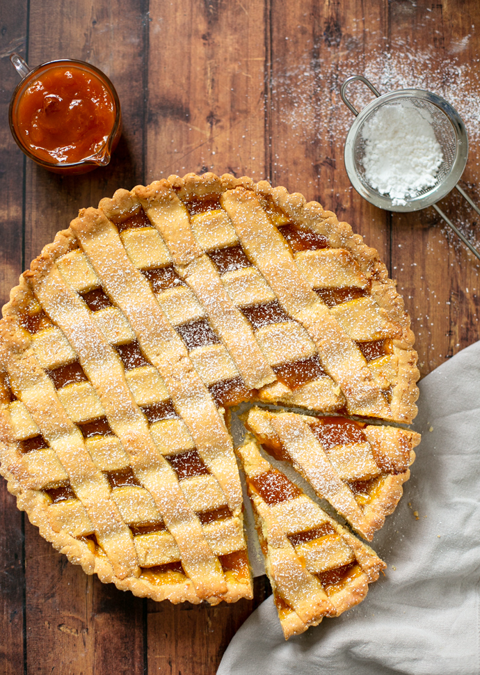 italian jam tart with 2 slices cut ready to be served. Confectioner sugar and peach jam on the side.