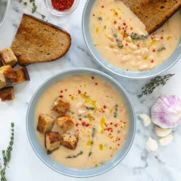 creamy cannellini soup served into two bowls and topped with croutons and grilled slice of bread, thyme leaves and red chili flakes.