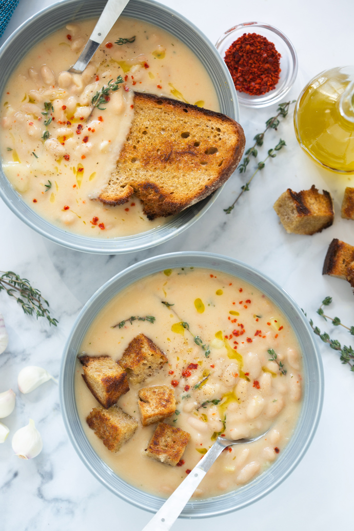 creamy cannellini soup served into two bowls and topped with croutons and grilled slice of bread, thyme leaves and red chili flakes.