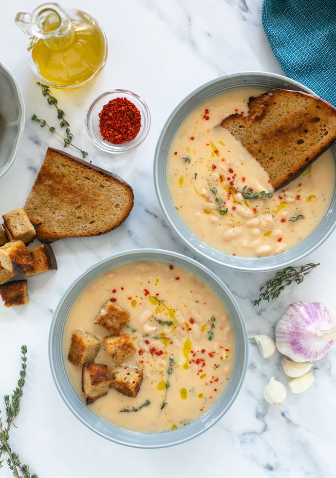 creamy cannellini soup served into two bowls and topped with croutons and grilled slice of bread, thyme leaves and red chili flakes.