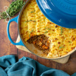 cottage pie in a cast iron casserole pot, topped with fresh thyme sprigs, and some mashed potatoes removed to show the minced beef beneath.