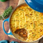 cottage pie in a cast iron casserole pot, topped with fresh thyme sprigs, and some potato mash removed to show the ground beef beneath. Image with graphic for Pinterest