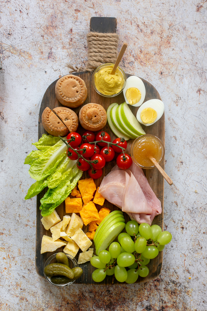 The Ploughman's lunch board featuring: pork pies, mustard, boiled eggs, cherry tomatoes, gem lettuce, cheddar, ham slices, grapes, gherkins, chutney.