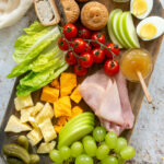 Ploughman's lunch board image showcasing pork pies, mustard, boiled eggs, sliced apple, gem lettuce leaves, ham slices, cheddar cheese, grapes and gherkins.
