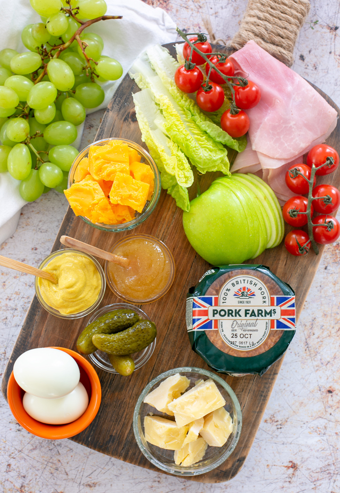 Recipe ingredients: grapes, ham slices, cherry tomatoes, cheddar cheese, pork pie, sliced green apple, boiled eggs, gherkins, mustard, chutney, gem lettuce.