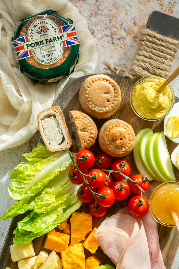 Ploughman's lunch close up showing pork pies, mustard, boiled eggs, sliced apple, gem lettuce leaves, ham slices, cheddar cheese.