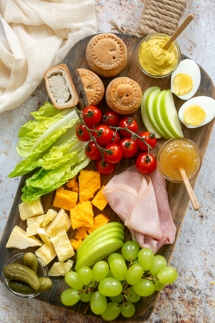 Ploughman's lunch board showcasing pork pies, mustard, boiled eggs, sliced apple, gem lettuce leaves, ham slices, cheddar cheese, grapes, gherkins.