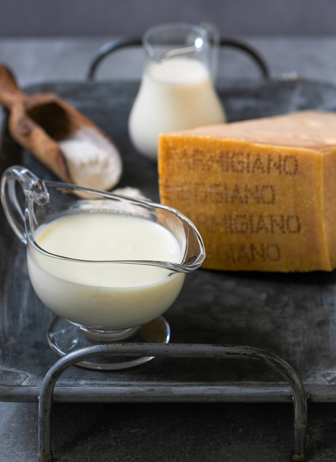 Parmigiano Reggiano white sauce in a saucer, next to Parmigiano Reggiano cheese wedge, flour and milk in the background.