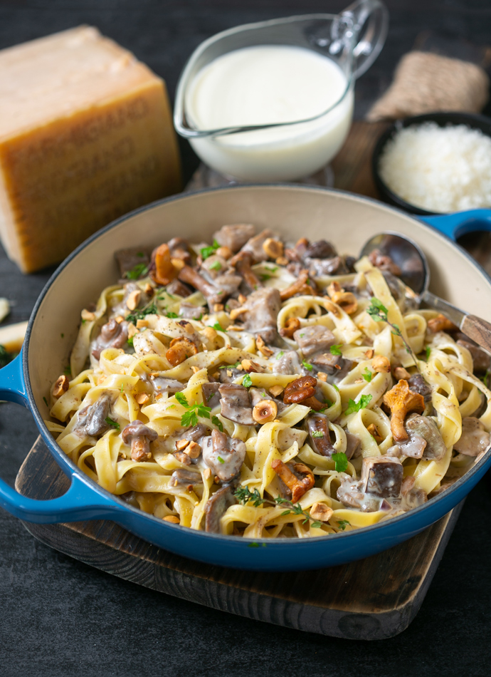 tagliatelle pasta with parmesan sauce, wild mushrooms and roasted hazelnuts, in a large cast iron shallow pot. Served next Parmigiano Reggiano cheese wedge and extra sauce on the side.