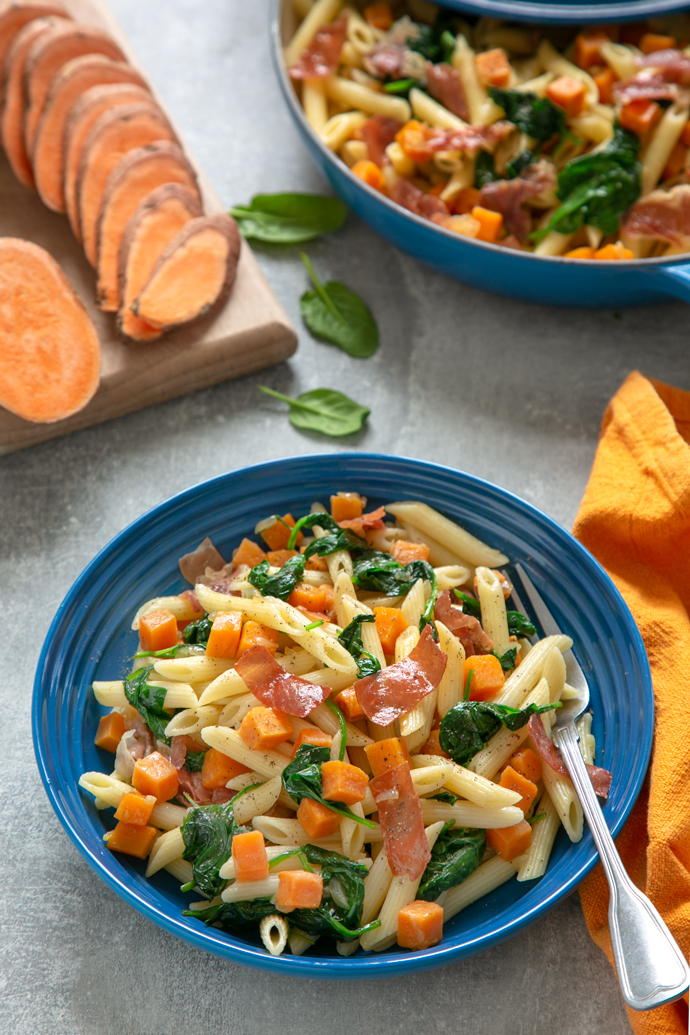 sweet potato penne pasta with crispy prosciutto adn sauteed spinach leaves served in a blue plate.