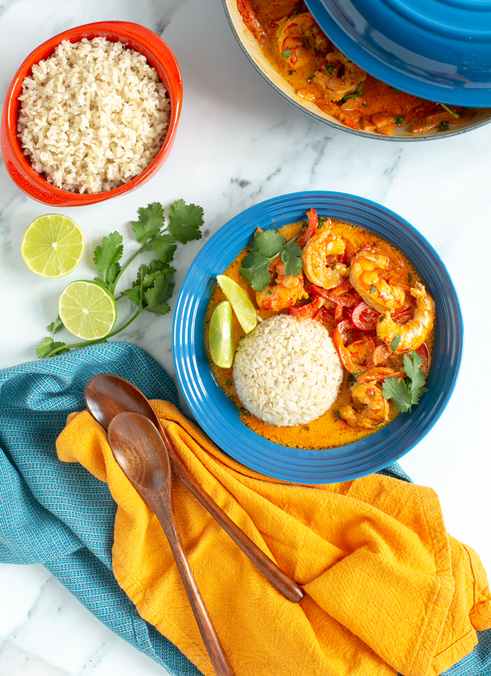 Prawn red curry served with rice, lime wedges and fresh coriander in a shallow plate, and extra brown rice on the side.