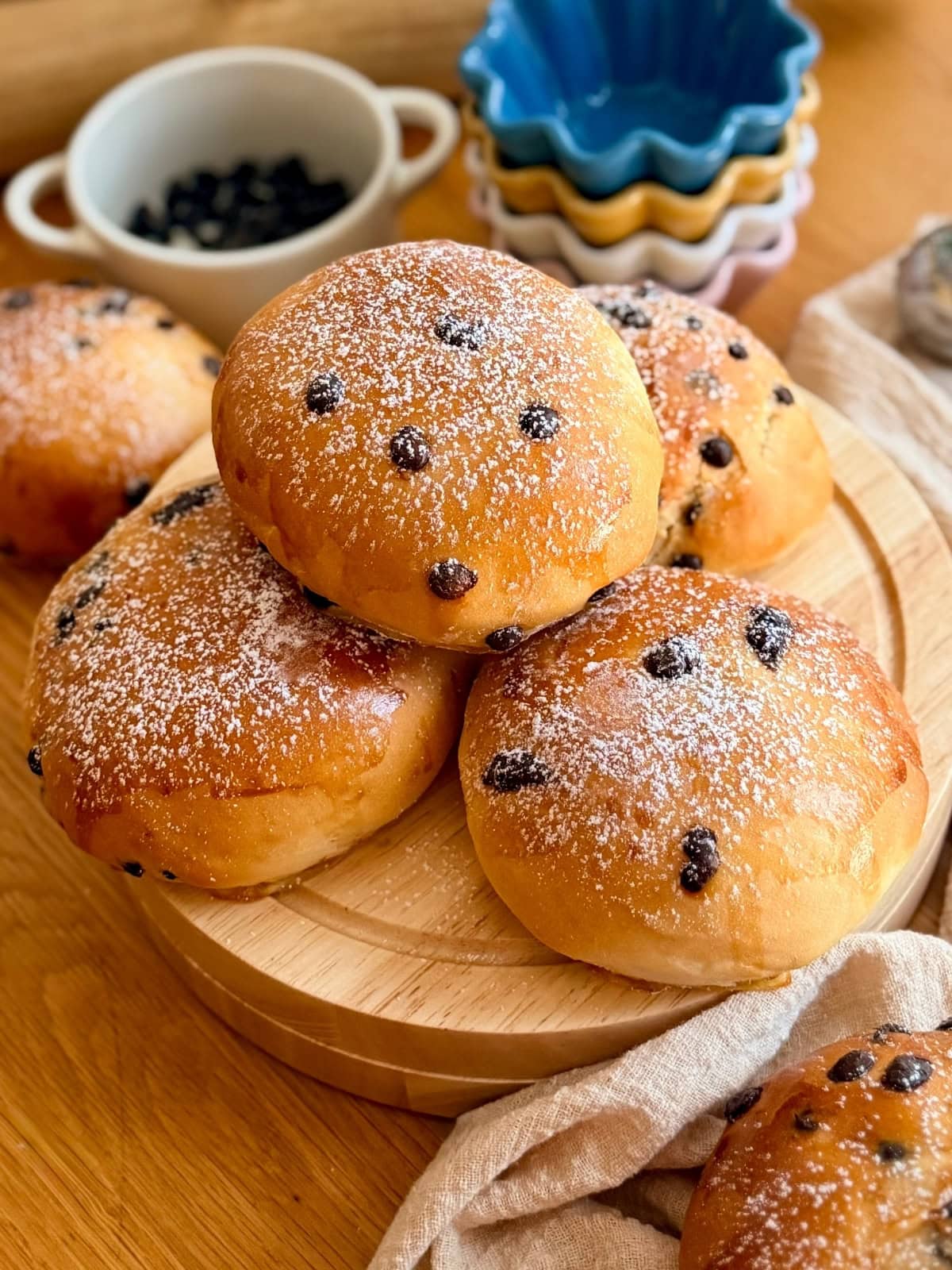 Golden milk buns topped with chocolate chips dusted with powdered sugar, stacked on a round wooden board