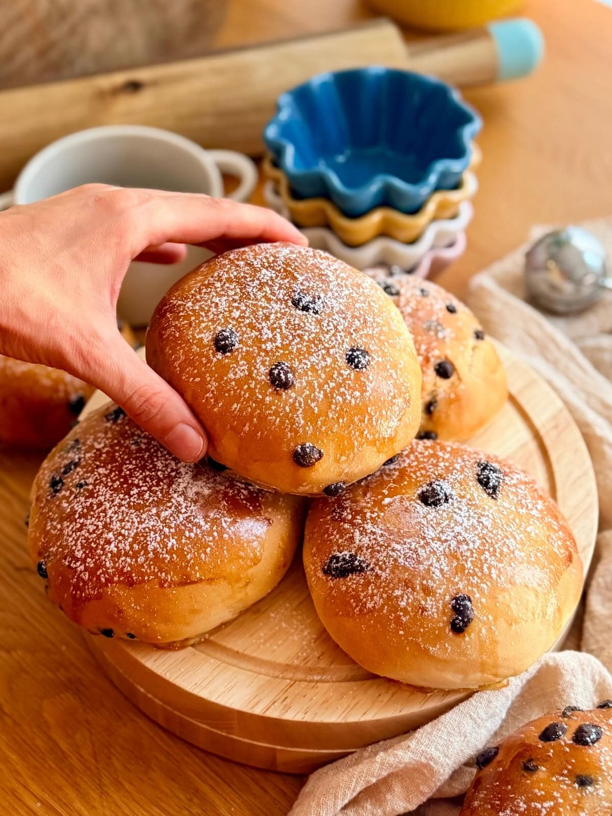 A hand picking up a chocolate chip milk bun from a pile of homemade pangoccioli on a wooden board